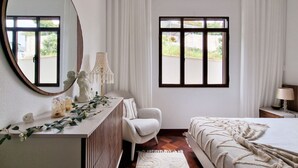 A bedroom with a large mirror, a bedside table with a lamp, a chair, and a bed with a blanket. - Villa Branca (Ponta do Sol)