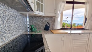A kitchen with granite countertops, white cabinets, a black stove, and a window with a view of greenery. - Villa Branca (Ponta do Sol)