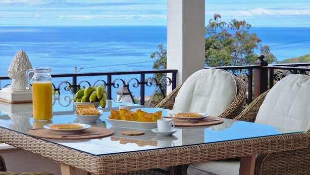 A table with breakfast items and a glass of orange juice, overlooking a scenic ocean view. - Villa Branca (Ponta do Sol)
