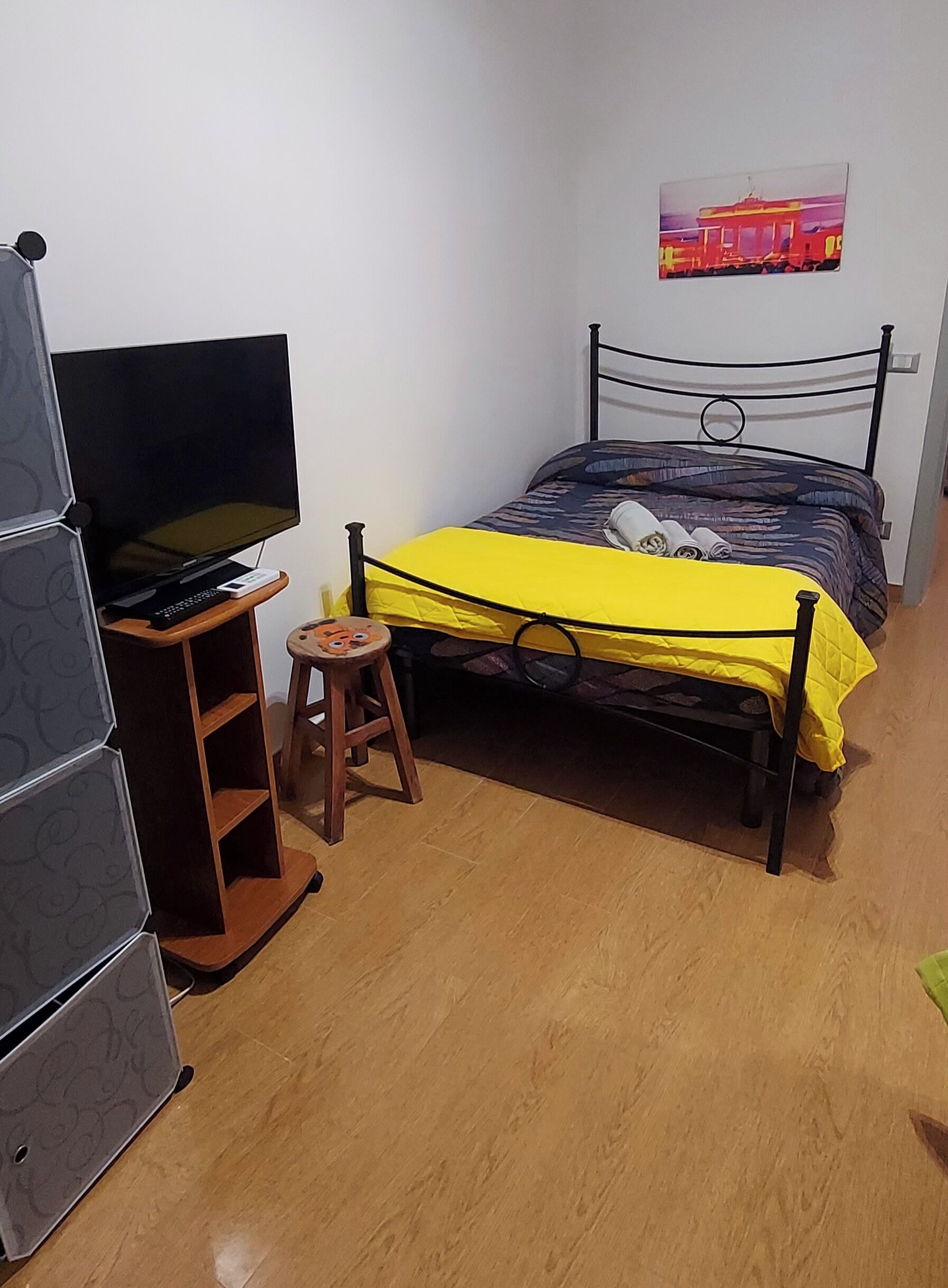 A bedroom with a bed, a TV on a wooden stand, a small stool, and a wall-mounted picture.