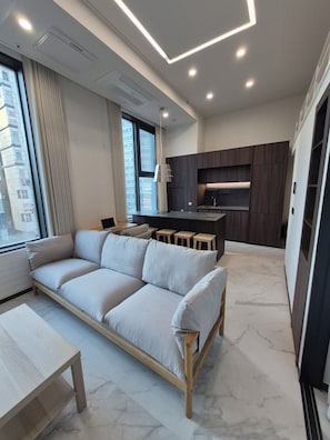 Relax in this stylish living area with a sofa against a window, a small table, and a kitchen in the background.