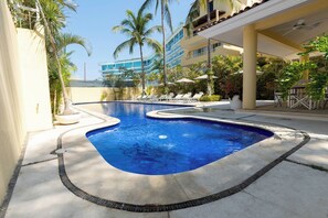 A swimming pool with a curved edge, surrounded by lounge chairs and palm trees, with a building in the background.