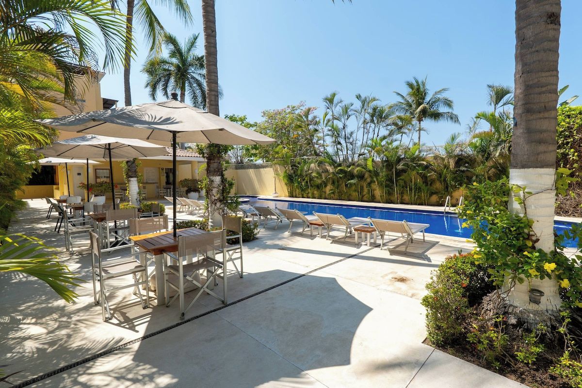 A poolside area with lounge chairs and umbrellas, surrounded by palm trees and a clear blue sky.