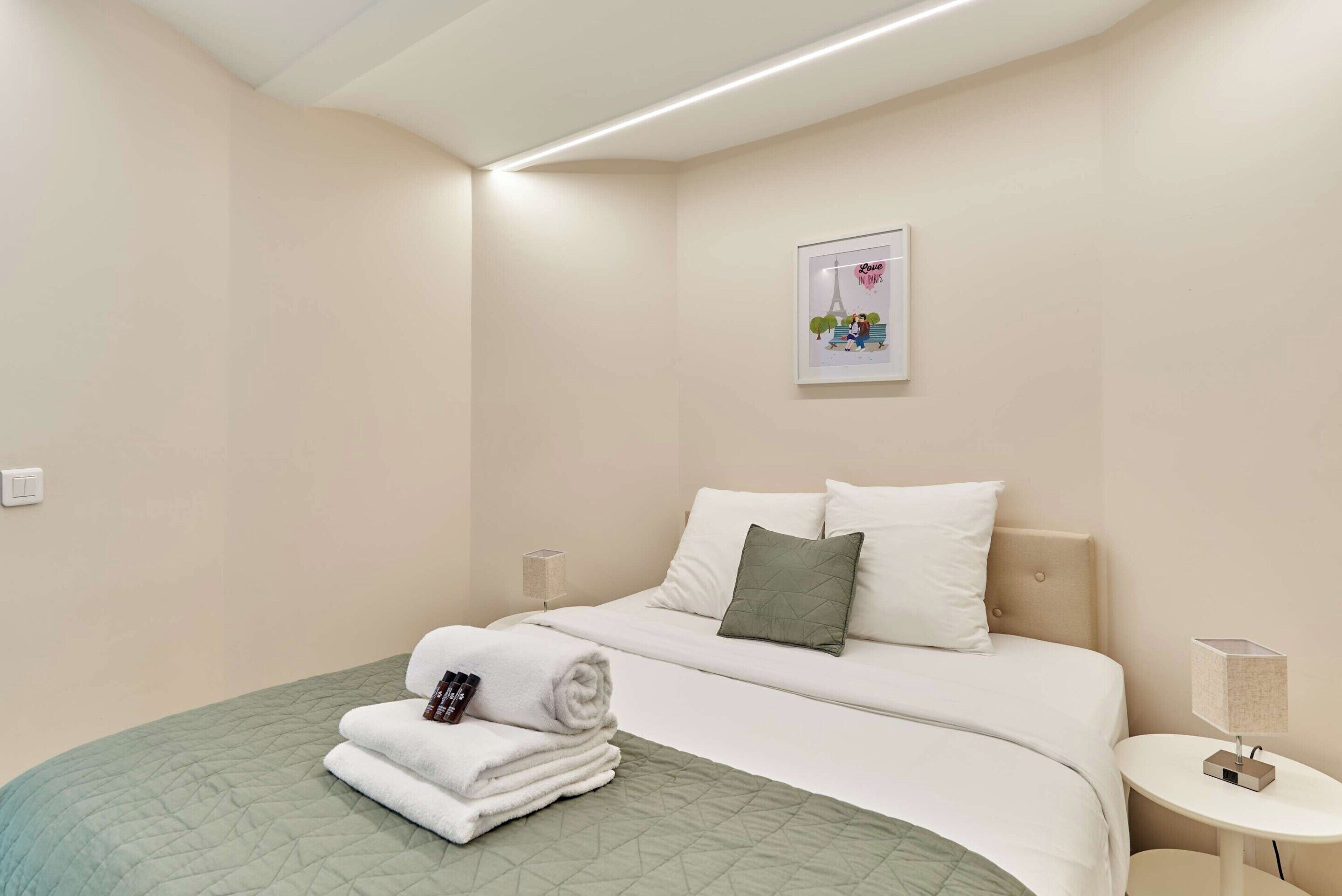 A neatly made bed with a green quilt, a pillow, and rolled towels. There is a framed picture on the wall and a bedside table with a lamp.