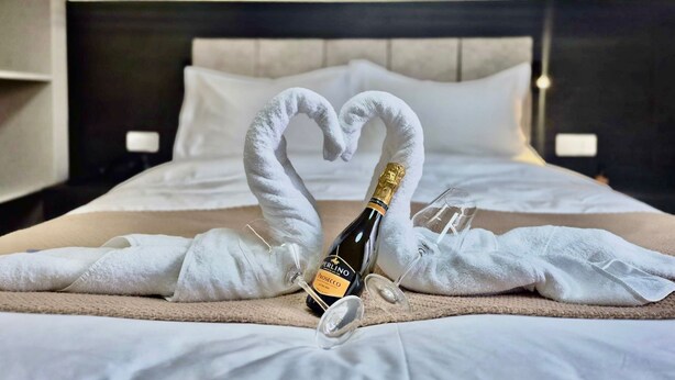 A bed with a heart-shaped towel arrangement, a bottle of Prosecco, and two wine glasses on a beige bedspread. - KIMMIY HOTEL (Belgrade)