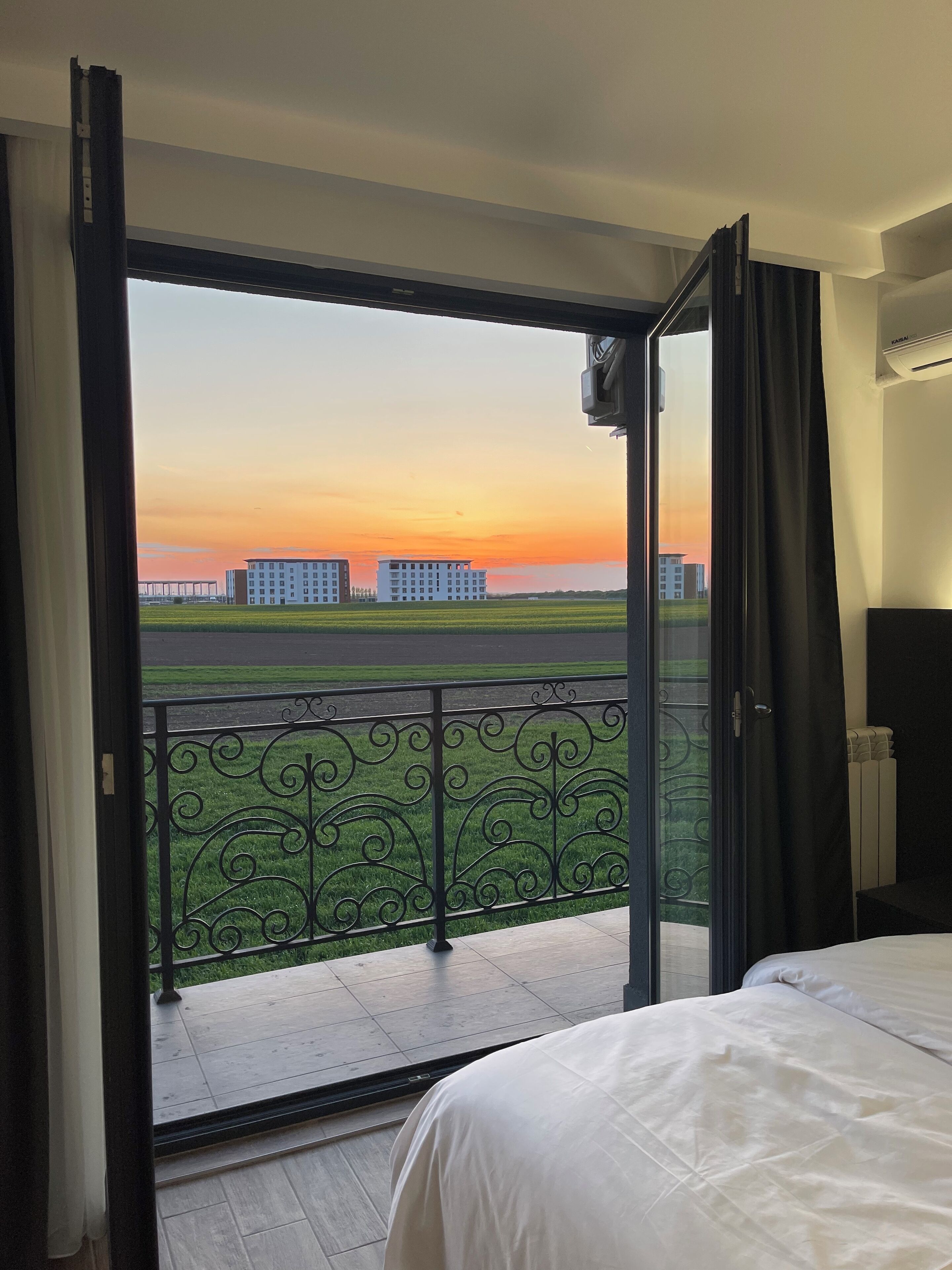 A hotel room with a balcony, a bed, and a view of buildings and a sunset.