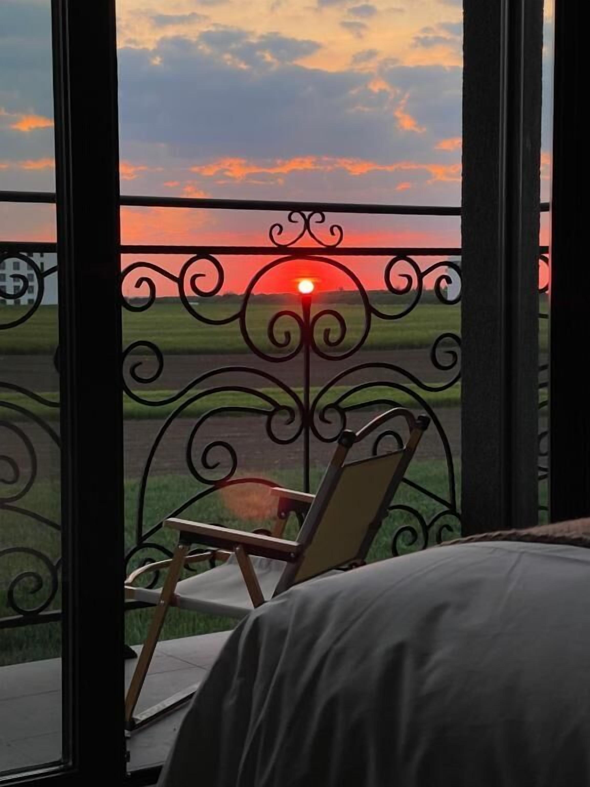 A balcony with a view of a sunset over a field, a chair, and a table.