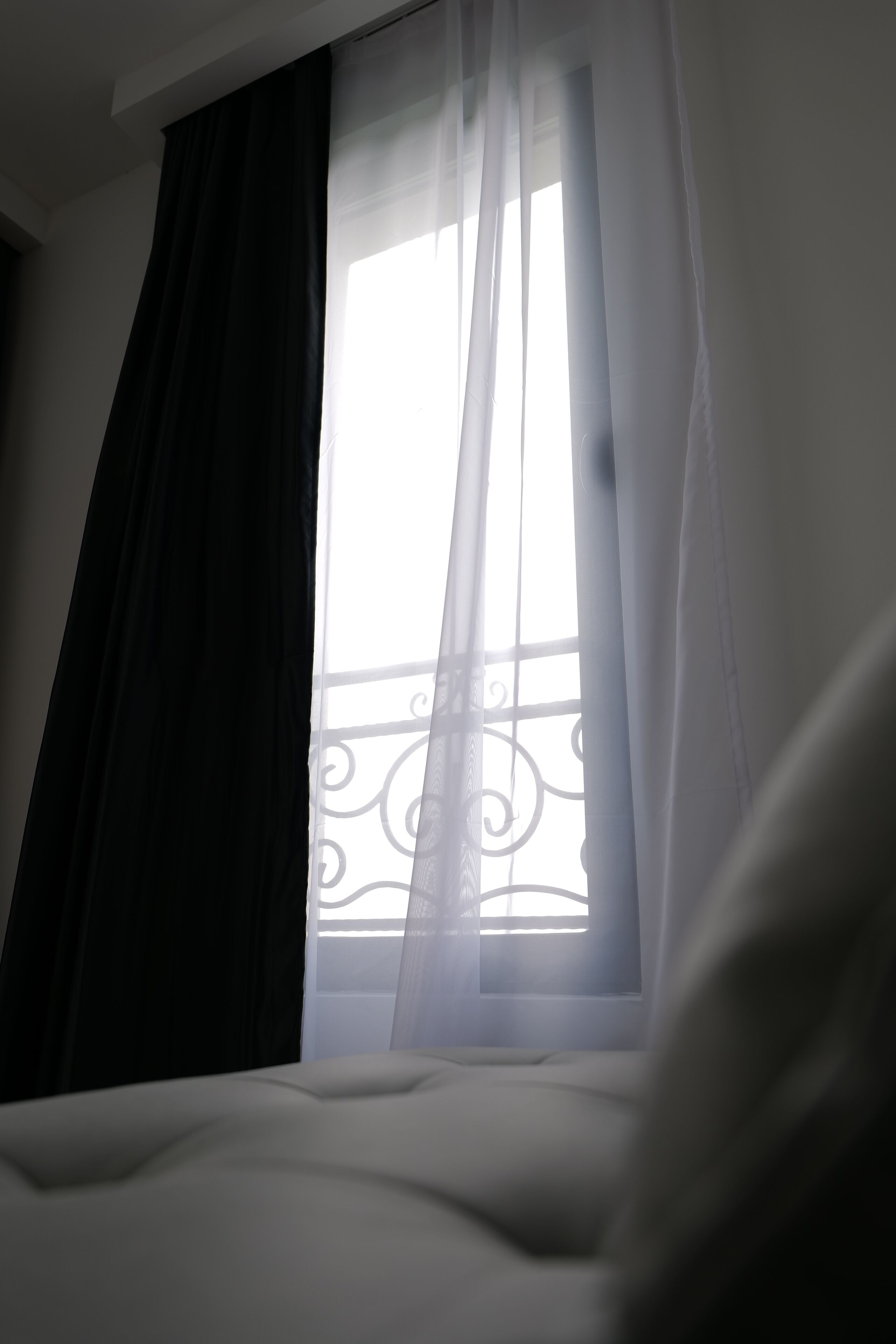 Una habitación con cama, ventana con cortinas traslúcidas y vista a un balcón con barandillas decorativas.