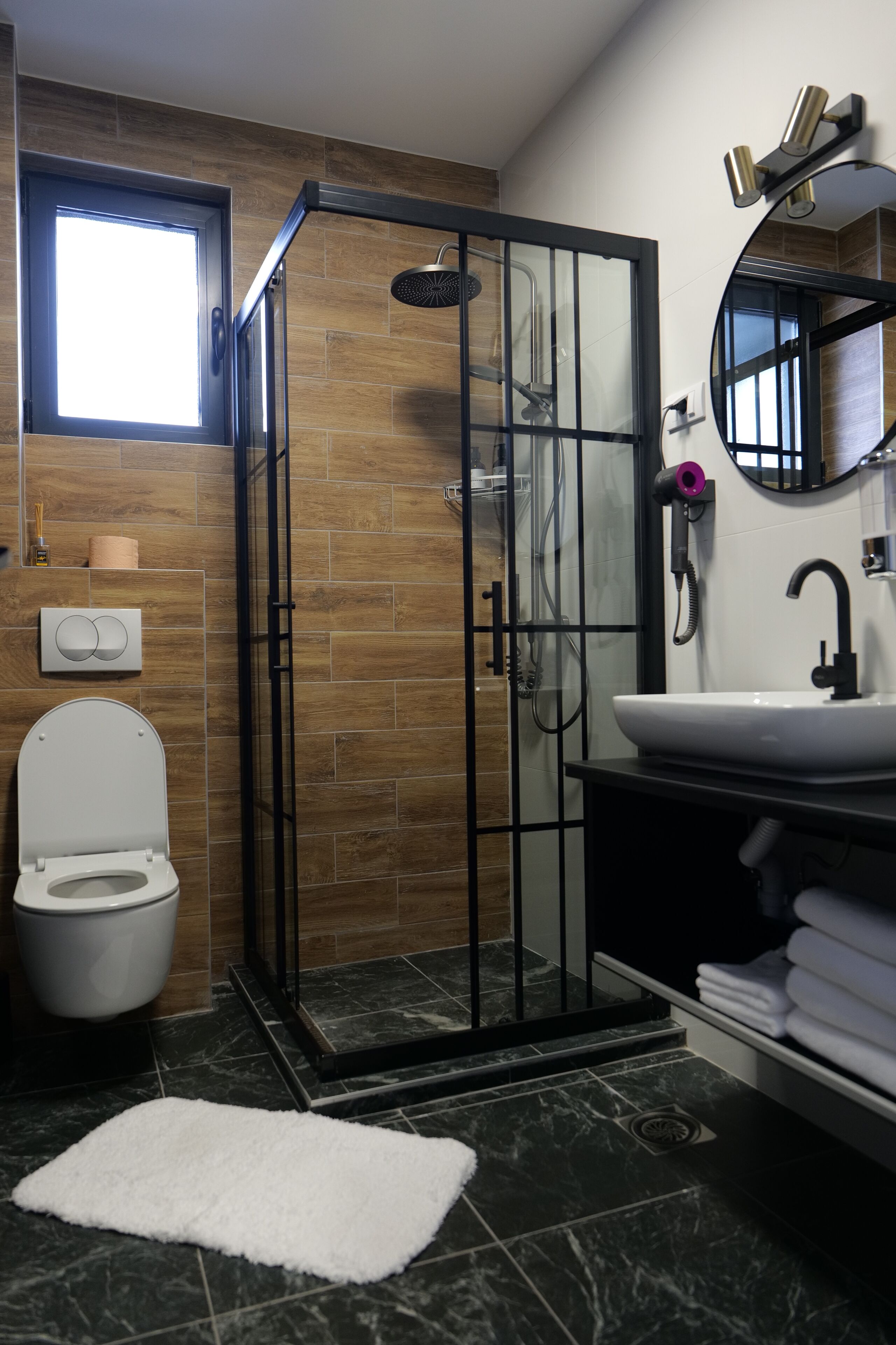 A modern bathroom with a glass-enclosed shower, a white toilet, a sink with a black countertop, and a white rug on the floor.