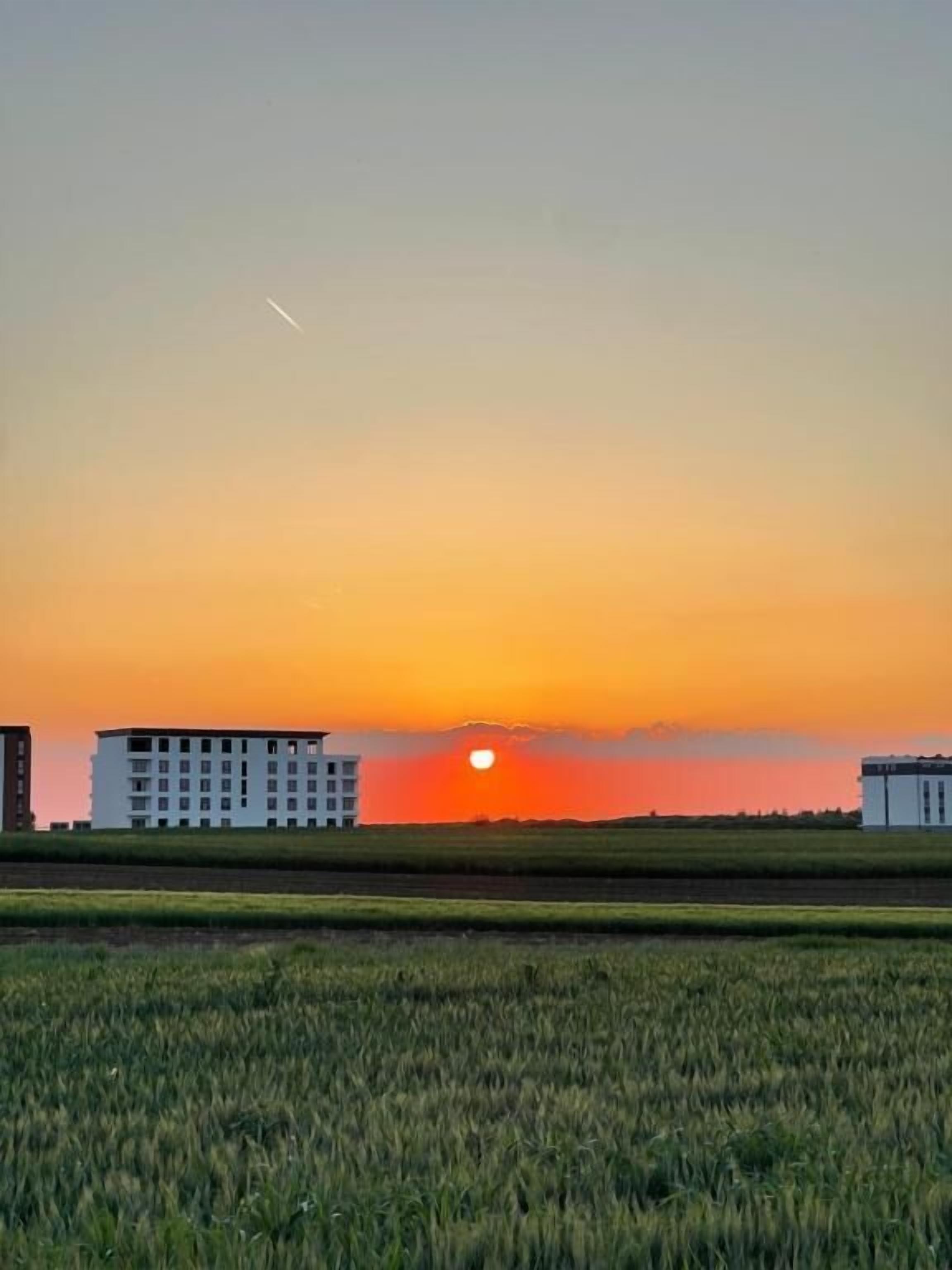 Ein Sonnenuntergang über einem Feld, im Hintergrund sind Gebäude zu sehen.