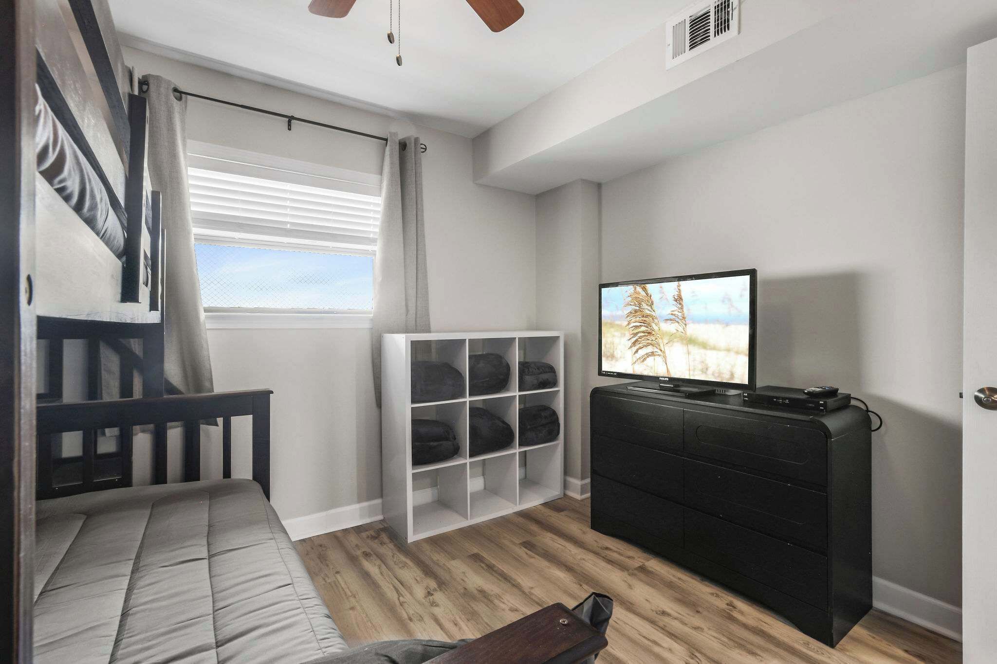 A room with a bed, a television, a storage unit, and a window with blinds.