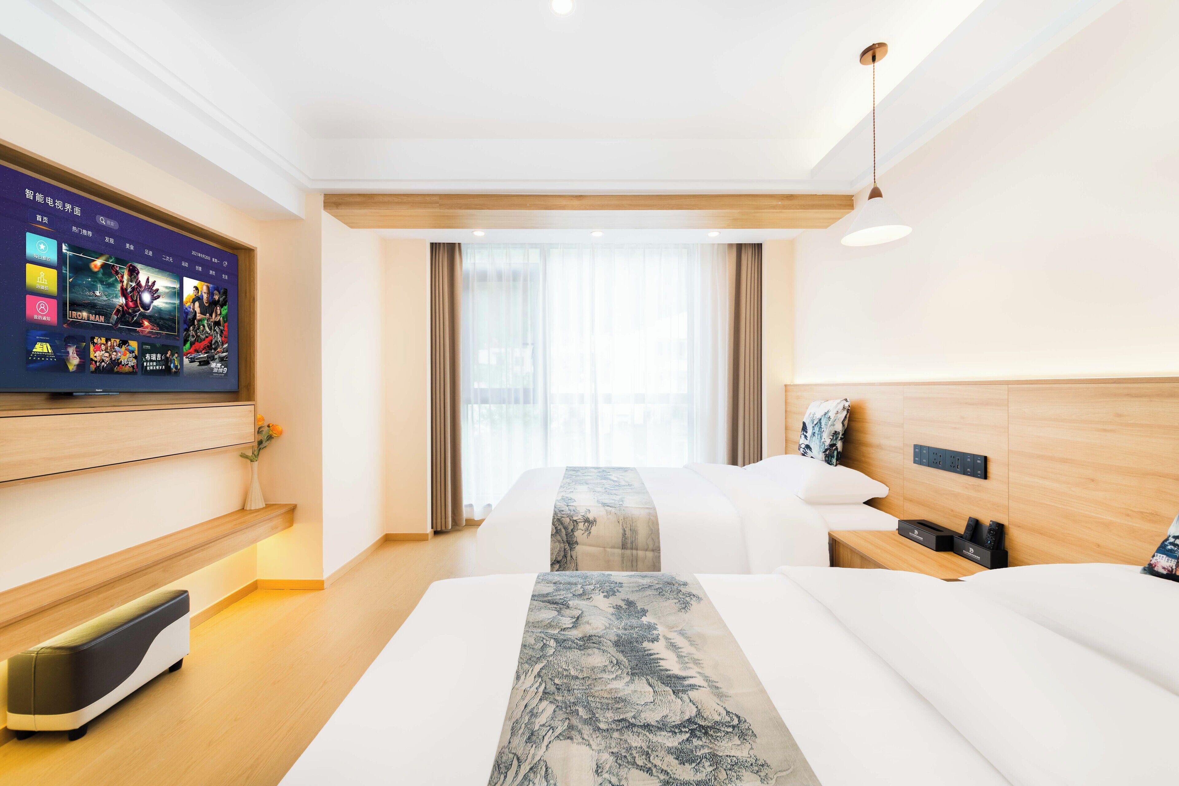 Hotellihuone, jossa on suuri, litteänäyttöinen televisio, jossa näkyy elokuvajulisteita, koristeellisella päätyosalla varustettu sänky, yöpöytä, jossa on puhelin ja laturi, sekä ikkuna, jossa on verhot.