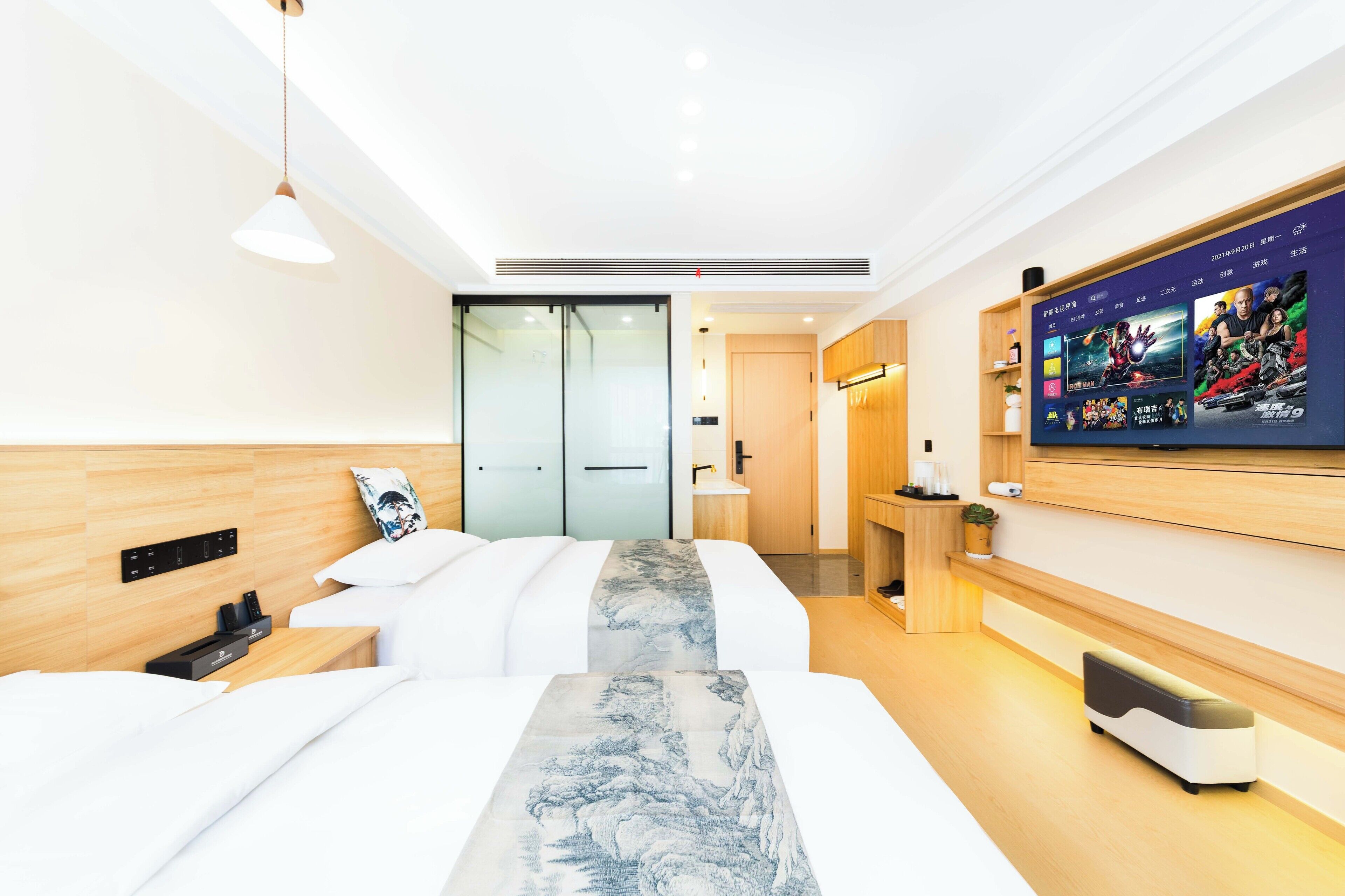 A modern hotel room with two beds, a TV mounted on the wall, and a small desk with a lamp.