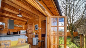 A cozy log cabin with a wooden interior, a kitchenette, a sofa with animal-themed cushions, and a large window overlooking a garden. - Pet-friendly Chalet in Surlingham, No Deposit (Norwich)