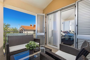 Een balkon met rieten meubels, een glazen tafel en uitzicht op een slaapkamer door een schuifdeur.