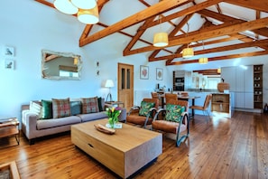 Een ruime woonkamer met houten balken, een bank, stoelen, een salontafel en een keuken op de achtergrond.