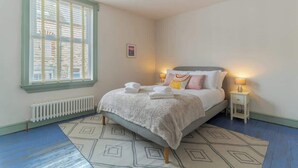 Ein Schlafzimmer mit Bett, Nachttischlampen, einem Fenster mit Jalousien, einem Heizkörper und einem gemusterten Teppich.