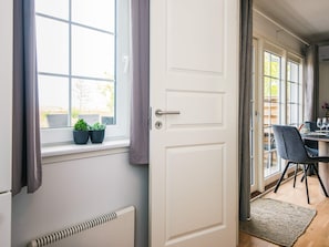 Una habitación con puerta blanca, ventana con cortinas, radiador, mesa con sillas y vista al exterior.