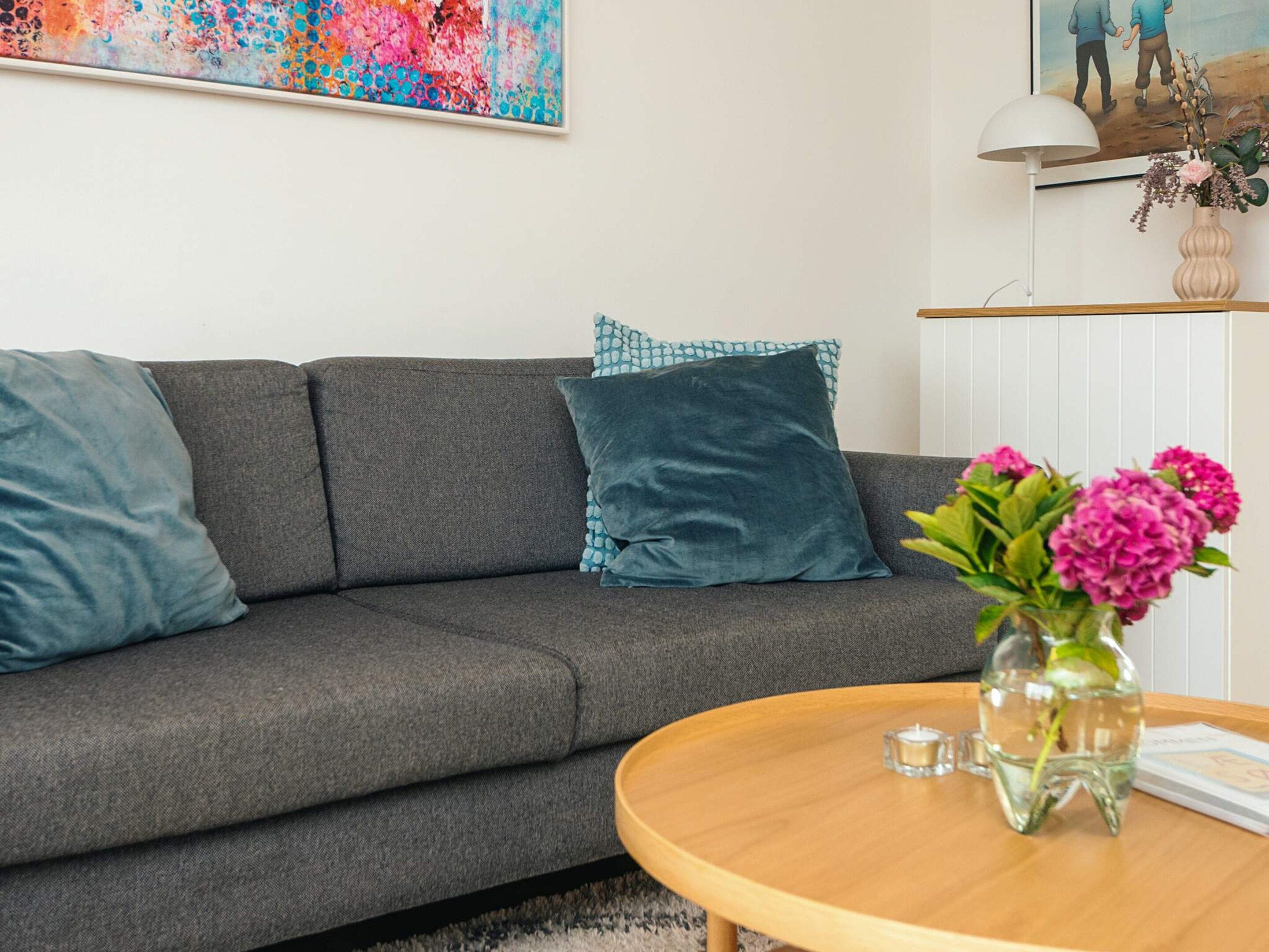 Una sala de estar moderna con un sofá gris, cojines turquesa, una mesa de centro de madera y un jarrón con flores rosas.