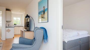 Una sala moderna con un sofá gris, una cama con ropa de cama gris, una zona de cocina con armarios blancos y un cuadro en la pared.