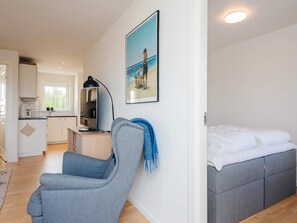 Una sala moderna con un sofá gris, una cama con ropa de cama gris, una zona de cocina con armarios blancos y un cuadro en la pared.