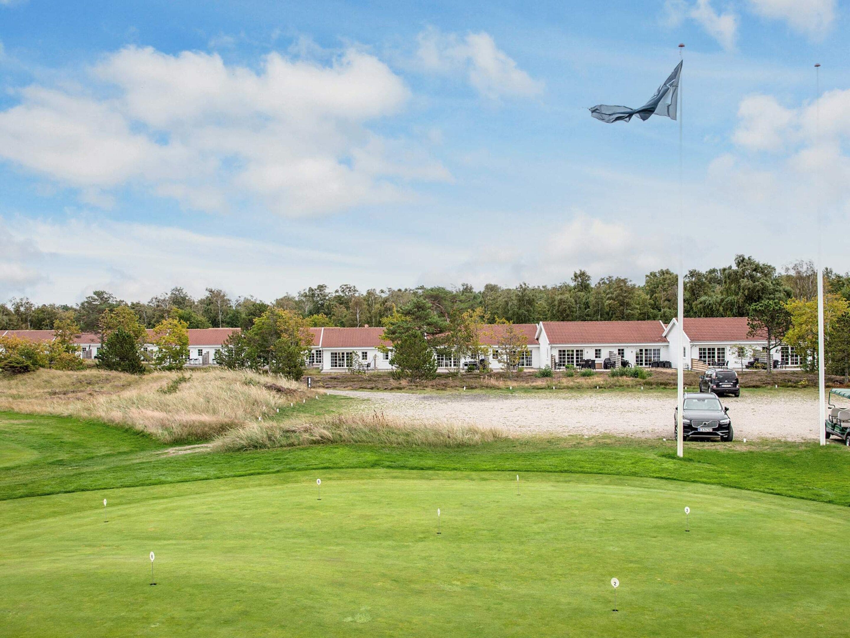 Un campo de golf con un mástil de bandera, un edificio con tejado rojo y un aparcamiento con coches.