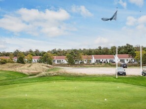 Un campo de golf con un mástil de bandera, un edificio con tejado rojo y un aparcamiento con coches.