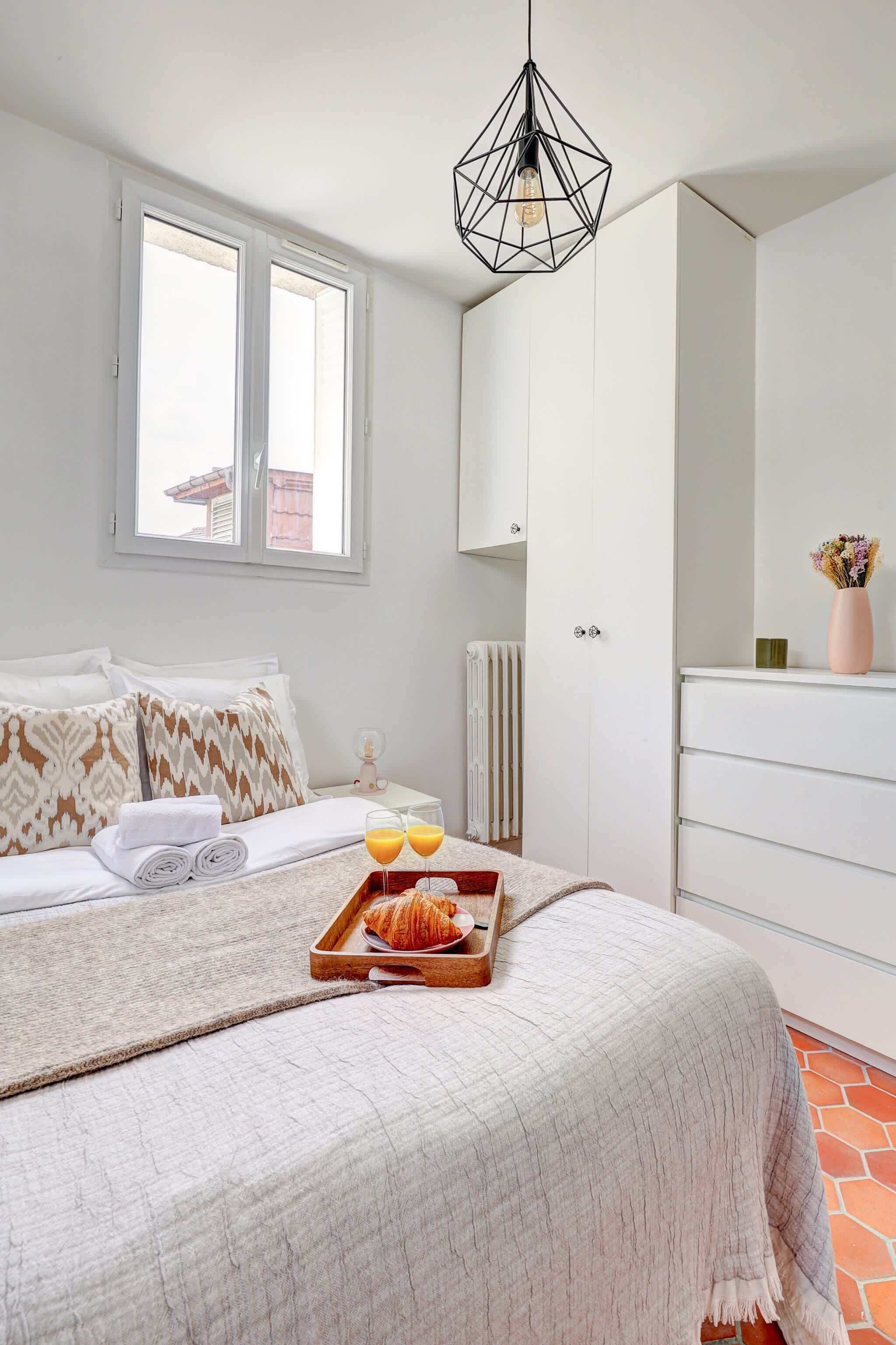 A neatly made bed with a tray of croissants and orange juice, a white wardrobe, and a geometric pendant light.