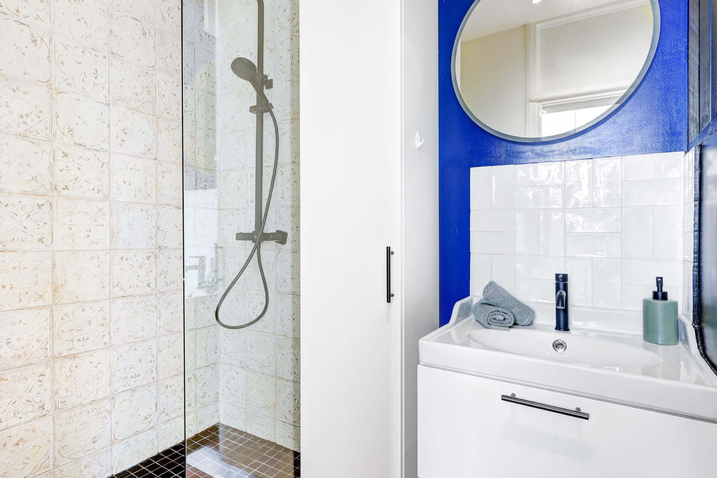 A bathroom with a glass shower enclosure, white tiles, a blue accent wall, a round mirror, a white sink, and a towel.
