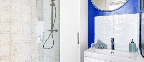 A bathroom with a glass shower enclosure, white tiles, a blue accent wall, a round mirror, a white sink, and a towel.