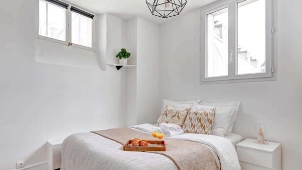 A bedroom with a bed, a bedside table, a nightstand, a window, and a wall-mounted shelf with a plant.