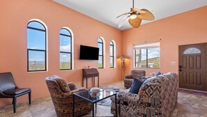 A spacious living room with a large sofa, armchairs, and a coffee table. There are large windows with arched tops, a flat-screen TV mounted on the wall, and a ceiling fan. - Foothills Casita | Summer Mountain Retreat (Oro Valley)