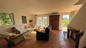 A spacious living room with a sofa, armchairs, a coffee table, and a fireplace. There is a large window providing natural light and a view of greenery outside.