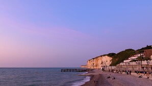 Ein Strand mit felsiger Küste, einer in das Wasser ragenden Mole und einer Klippe mit Gebäuden darauf.