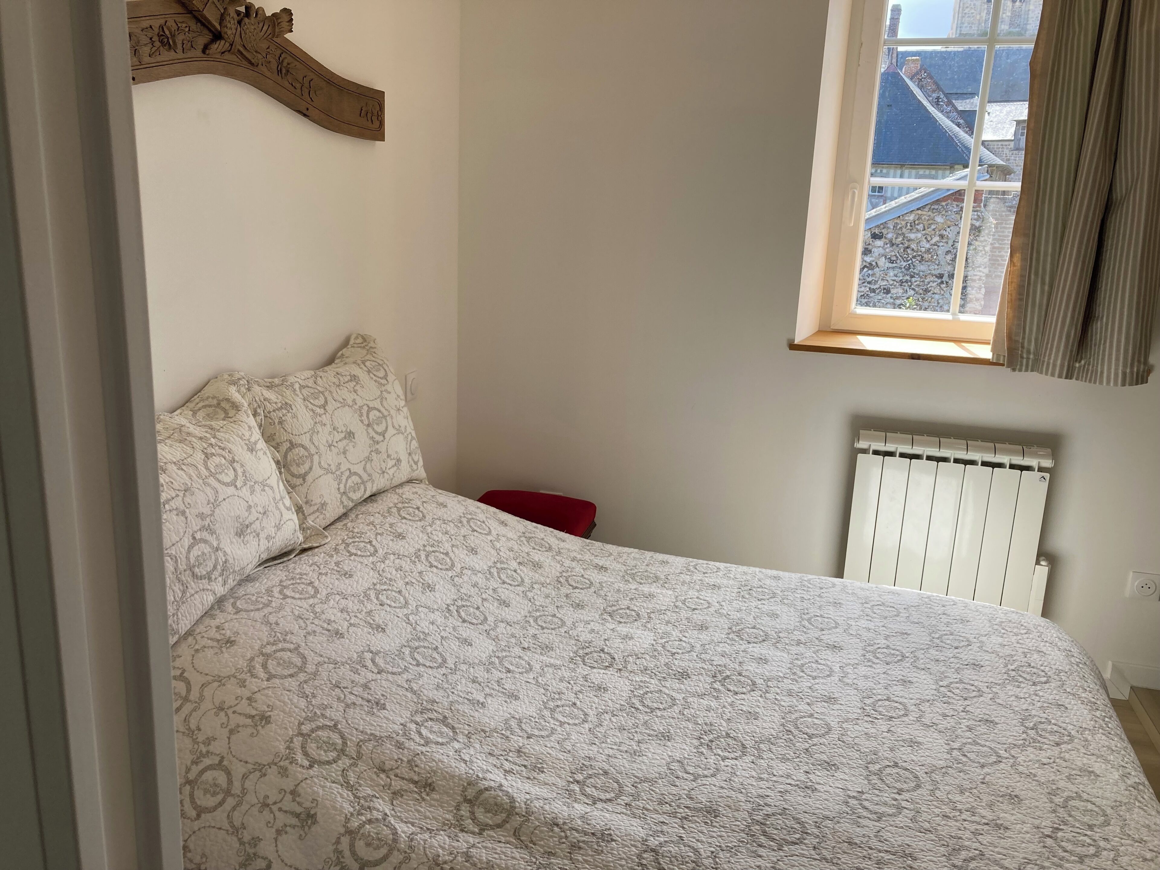 Et soveværelse med et mønstret sengetæppe, en rød stol, en radiator og et vindue med udsigt til bygninger.