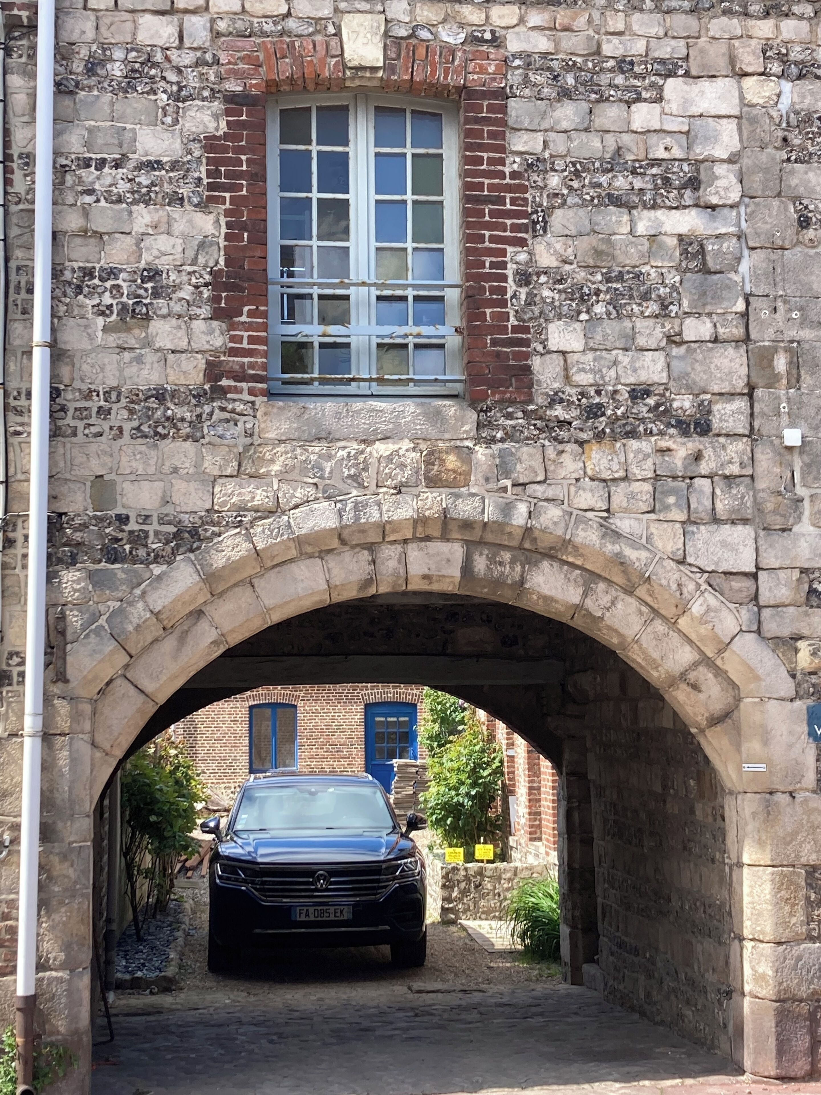 Ein steiner Torbogen führt zu einem Gebäude mit einem blauen Fenster, darunter ist ein schwarzes Auto geparkt.