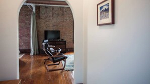 Una habitación con pared de ladrillo, un televisor sobre un mueble de madera, un sillón de cuero y un cuadro enmarcado en la pared.