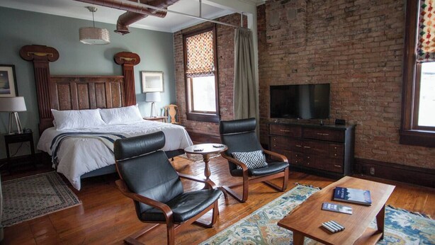 Habitación de hotel con una cama grande, dos sillones de cuero, una mesita redonda, televisor y pared de ladrillo.