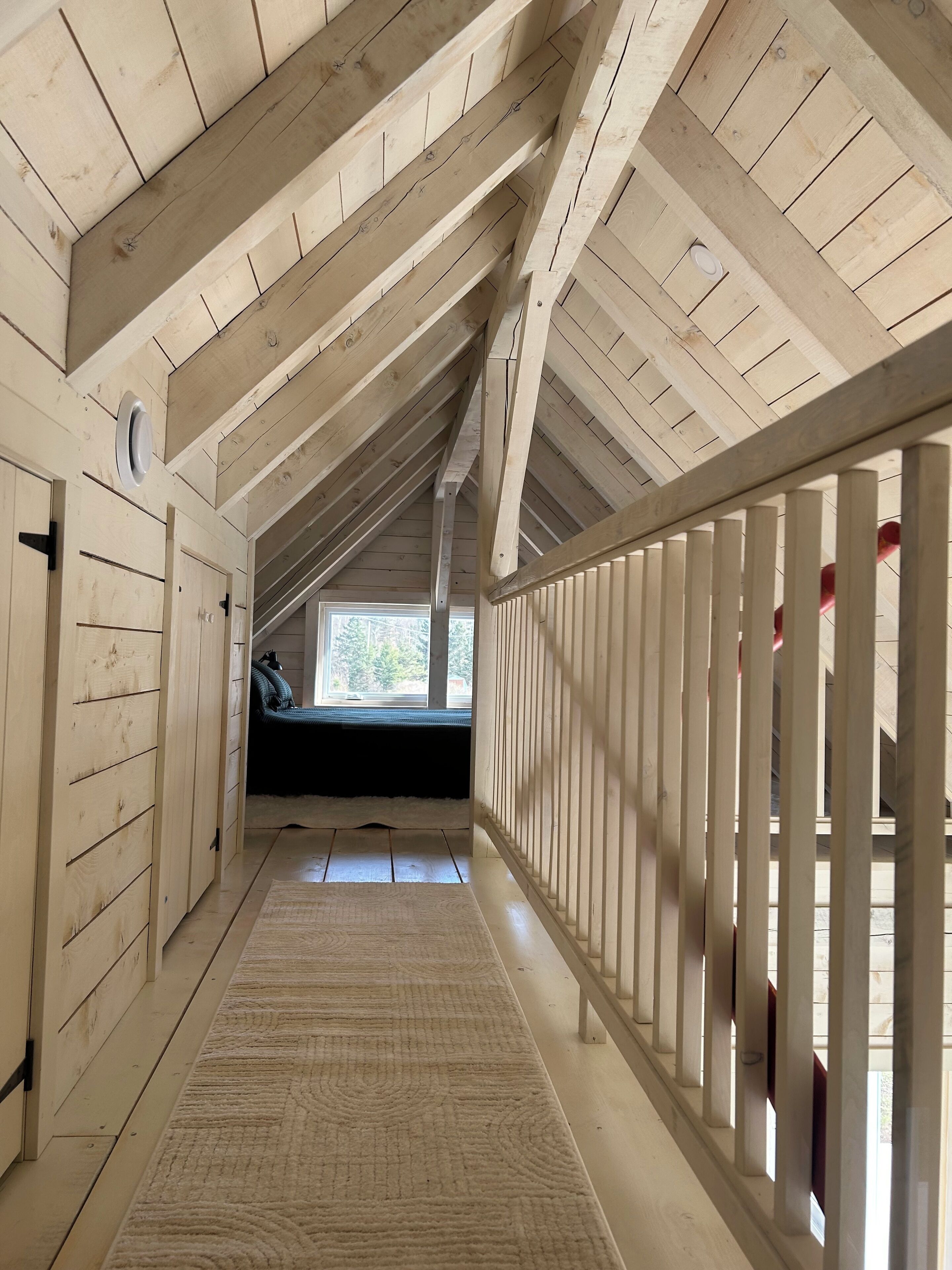 A narrow, wooden hallway with a sloped ceiling, a view to a window, and a white railing on one side.