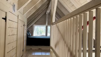 A narrow, wooden hallway with a sloped ceiling, a view to a window, and a white railing on one side.