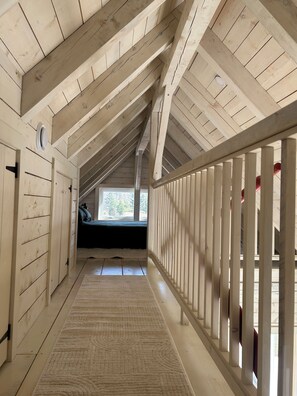 Un estrecho pasillo de madera con techo inclinado, una ventana al fondo y una barandilla blanca a un lado.