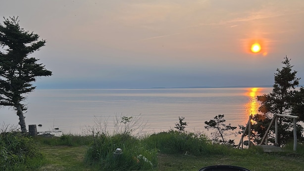 Ett lugnt utomhusområde med sittplatser och utsikt över solnedgången över ett stilla vatten, omgivet av grönska och en liten eldstad.