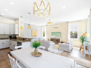 Cozinha moderna com armários brancos, bancada de granito e uma ilha central. Há uma área de jantar com mesa e cadeiras brancas, e uma sala de estar com sofá e televisão.