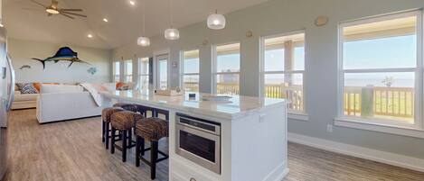 A modern kitchen with a central island, bar stools, and large windows offering views of the ocean.