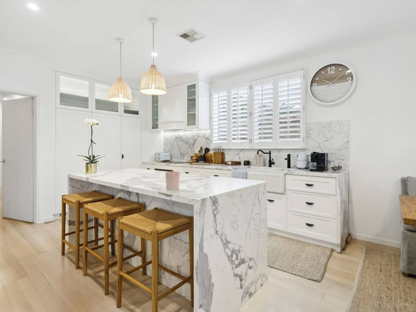 Une cuisine moderne avec un îlot en marbre, des tabourets en bois, des luminaires suspendus et une horloge au mur.