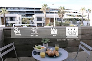 Una terraza con una mesa lista para el desayuno, vistas de un edificio moderno e íconos de servicios que incluyen estacionamiento, acceso a la playa y WiFi.