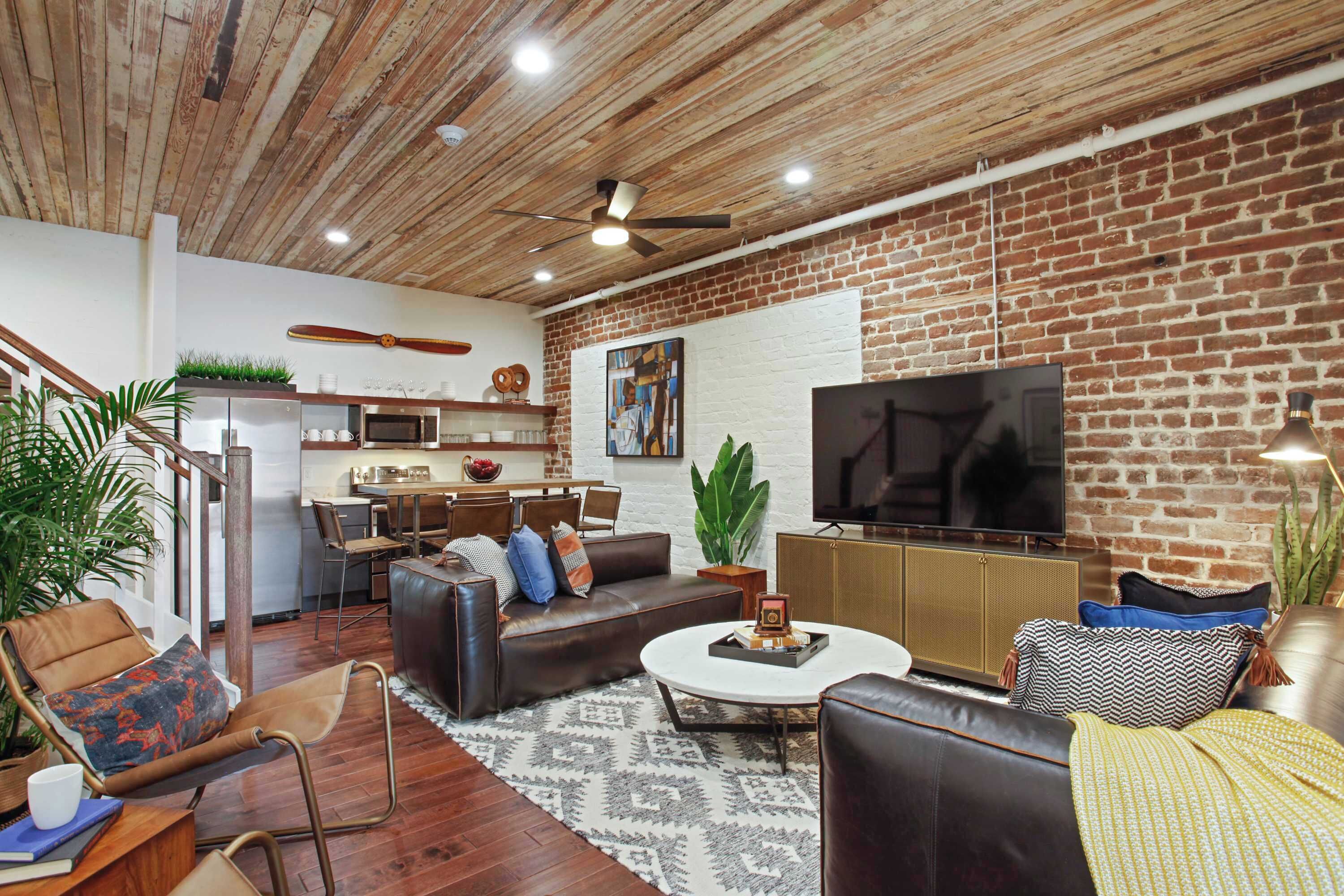 A modern living room with a brick wall, a flat-screen TV, a sofa, a coffee table, and a dining area with a kitchen in the background.