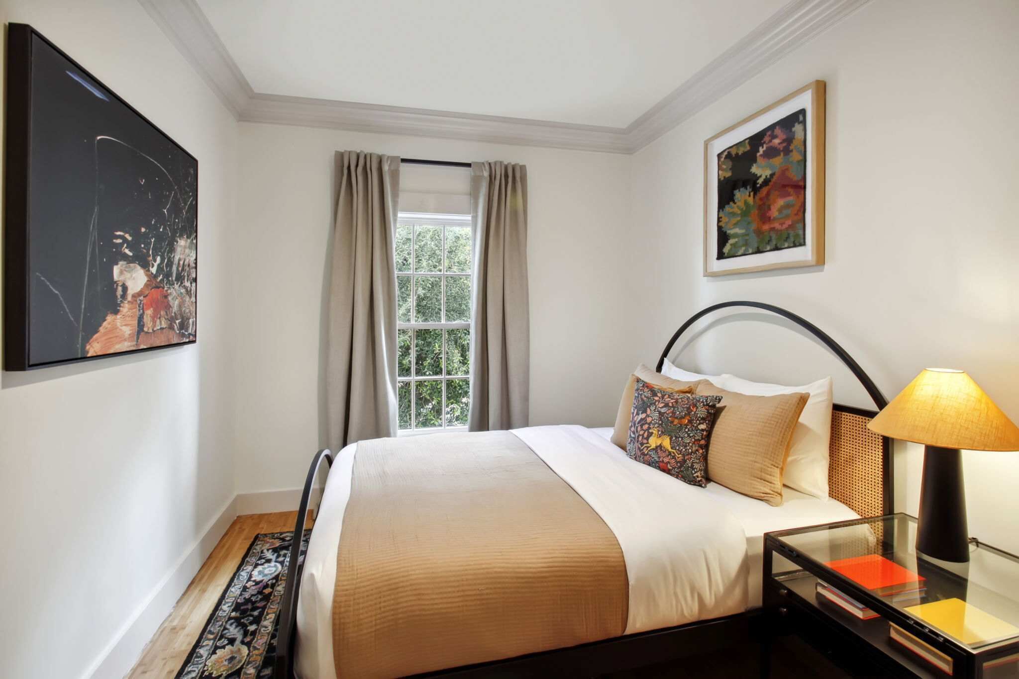 A hotel room with a bed, a nightstand, a lamp, a window with curtains, and a painting on the wall.