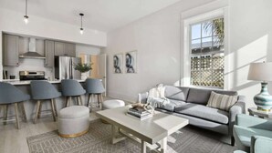 Un espace de vie moderne comprenant une cuisine, des chaises recouvertes d’un tissu gris, un canapé gris et un tapis à motifs.