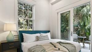 Une chambre à coucher comprenant un lit, une table de chevet, une lampe et une vue sur un jardin, accessible par une porte coulissante.